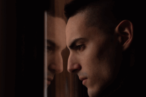 Young man staring out a window, his reflection visible in the glass, suggesting introspection, isolation, or waiting.