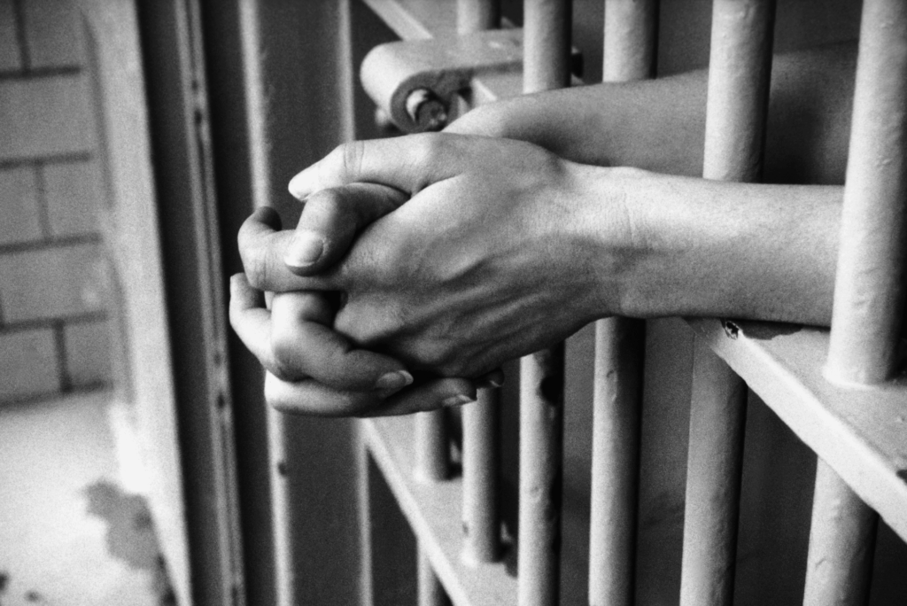 Black-and-white photo of a person’s hands clasped together, resting through the bars of a prison cell, evoking a sense of waiting, confinement, or contemplation.