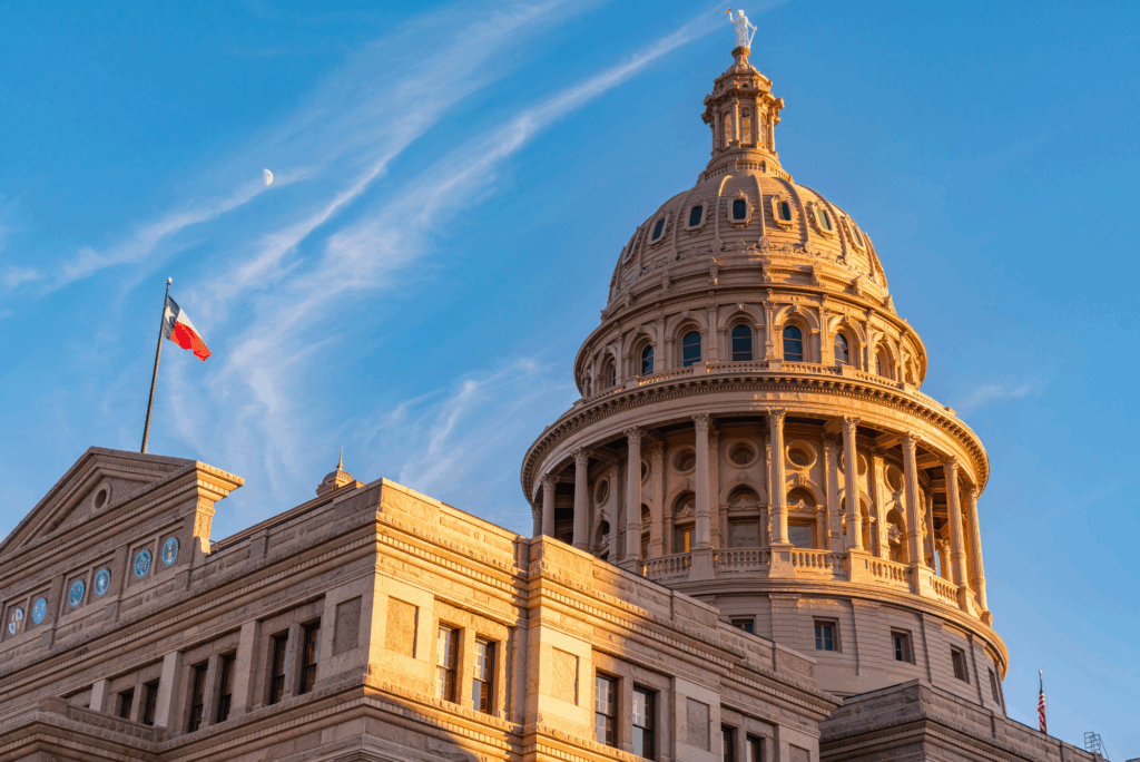 Exterior of the Texas State Capitol building in Austin, lit by warm sunlight under a blue sky with wispy clouds and a visible half-moon. The Texas flag is raised on a flagpole.