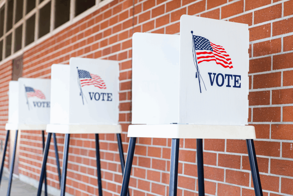Row of voting booths lined up against a red brick wall, each marked with an American flag and the word "VOTE" in bold blue letters.